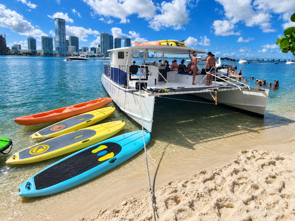Catamaran docked on beach with colorful paddleboards and city skyline.