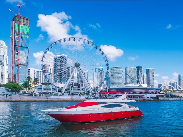 Red and white yacht on water with city skyline and Ferris wheel in background.