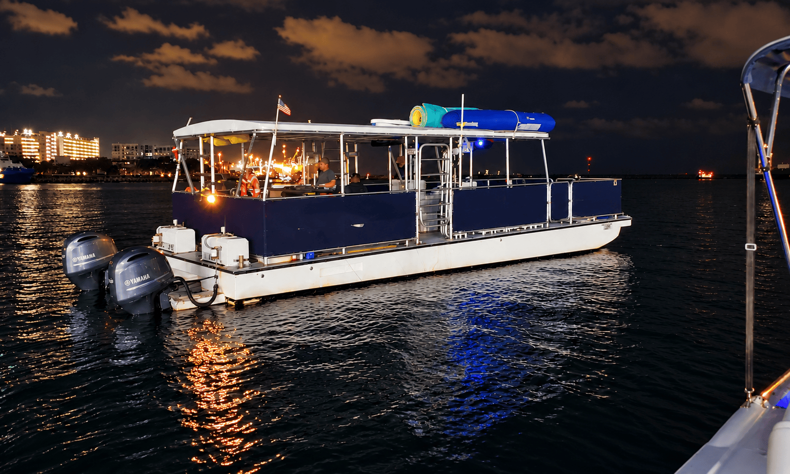 A pontoon boat with lights on water at night near cityscape.