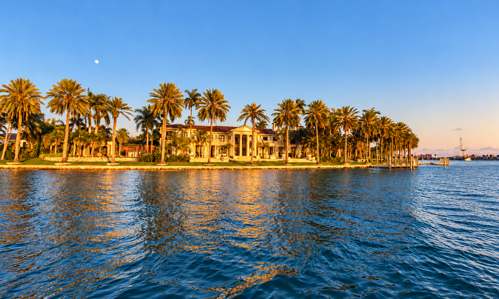 Luxurious mansion with palm trees by a waterfront at sunset.